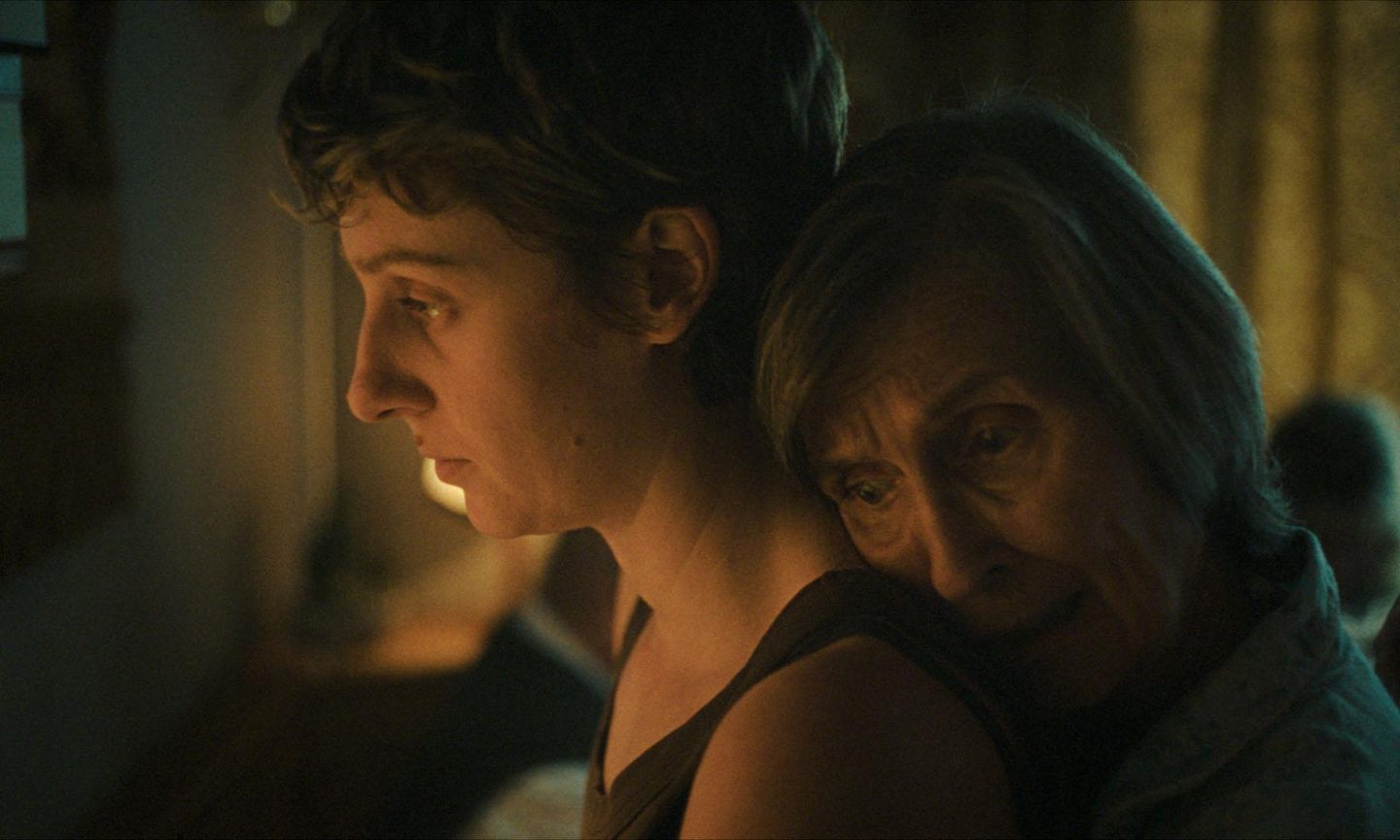 A young woman looks ahead in warm lamplight while an older woman rests her head against her shoulder