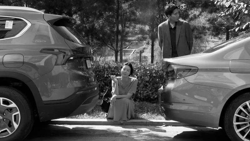 In black and white, a woman in a loose dress crouches on a kerb between two parked cars, one hand raised, while a man in a jacket stands behind a hedge looking away